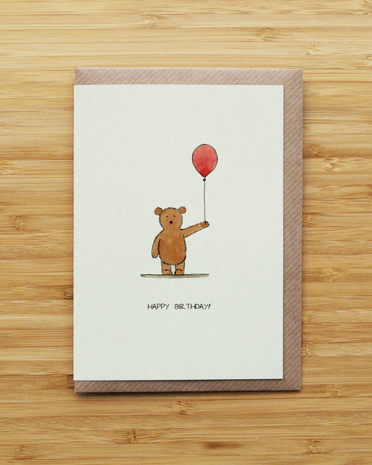 A birthday card with a brown envelope, featuring a watercolour illustration of a brown bear holding a red balloon with the text 'HAPPY BIRTHDAY!!' below, displayed on a light wood background.