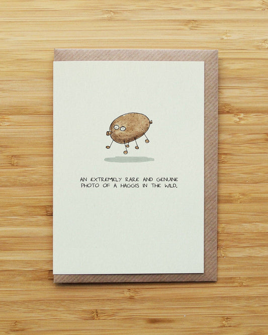 A greeting card with a brown patterned border and a small cartoon haggis character with legs, placed on a light wood background. The card reads, 'AN EXTREMELY RARE AND GENUINE PHOTO OF A HAGOS IN THE WILD.