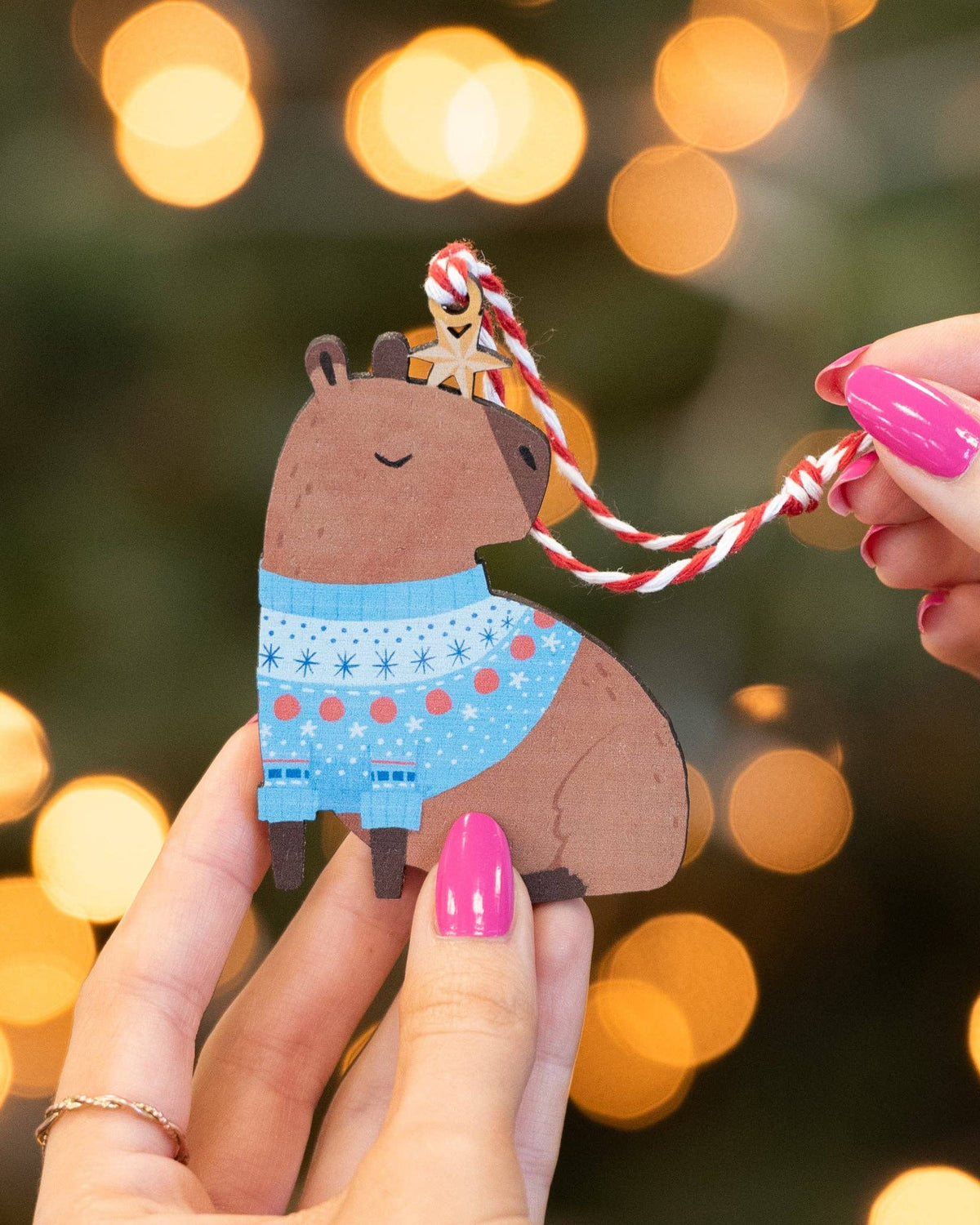 A small wooden capybara ornament with a blue patterned sweater and a red-and-white hanging string, held in two hands against a blurred festive background with warm bokeh lights.