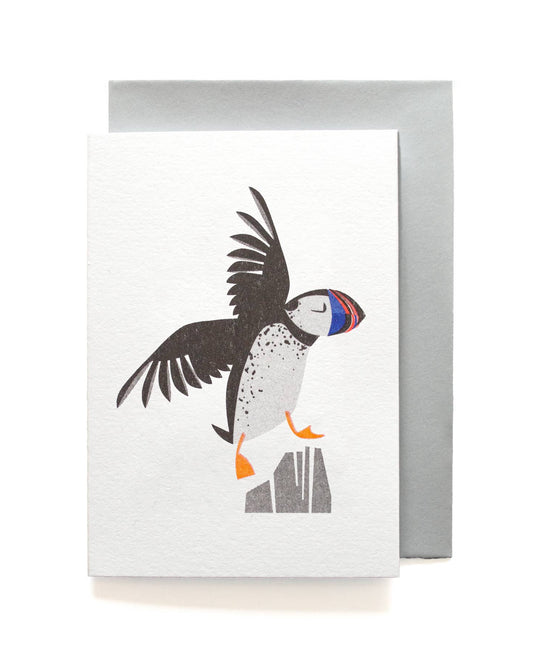 A white textured greeting card featuring a stylized puffin illustration in black, white, gray, orange, and blue, shown with a silver-gray envelope behind it on a white background.
