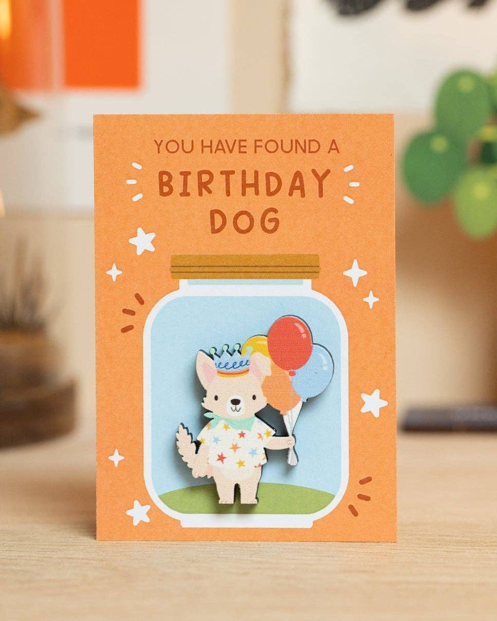 An orange backing card with a cute dog with balloons pin illustrated to look like it’s in a jar, standing on a wooden surface. The card reads 'YOU HAVE FOUND A BIRTHDAY DOG' and is photographed in a styled indoor setting with blurred decor in the background.