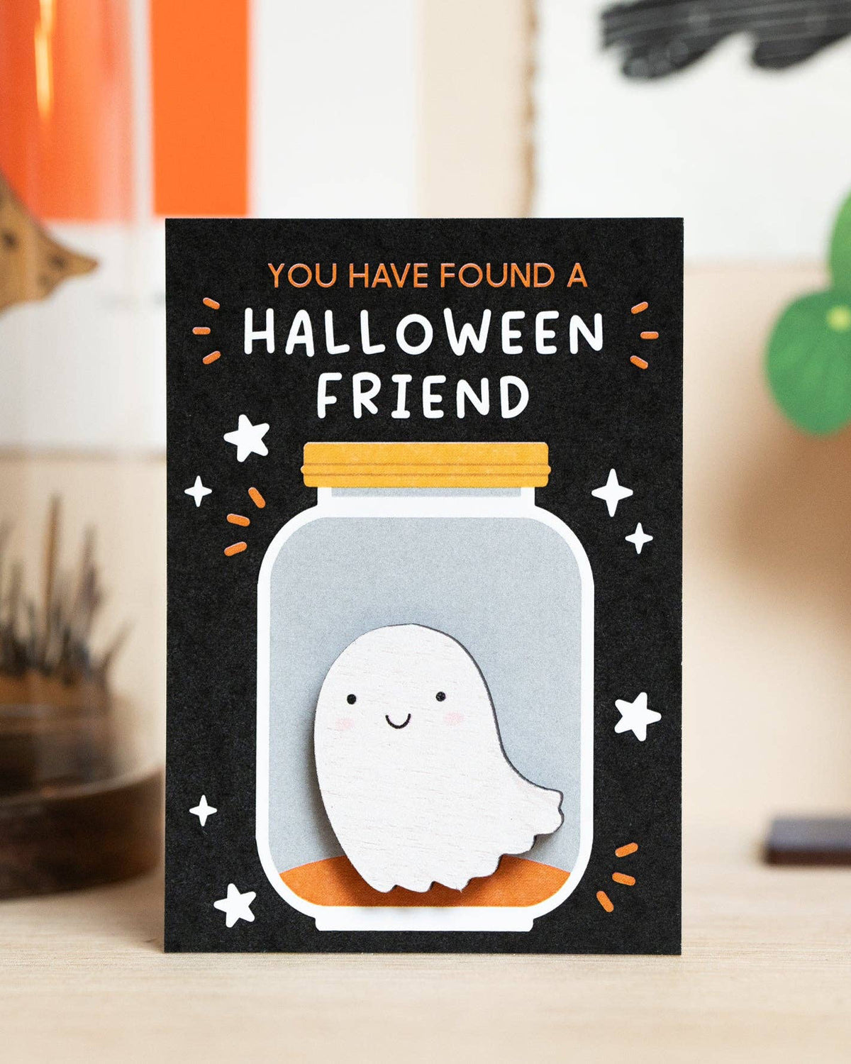 A Halloween-themed ghost pin on backing card standing upright on a tabletop. The card has a black background with the text 'YOU HAVE FOUND A HALLOWEEN FRIEND' above a jar illustration, with the white ghost pin attached.
