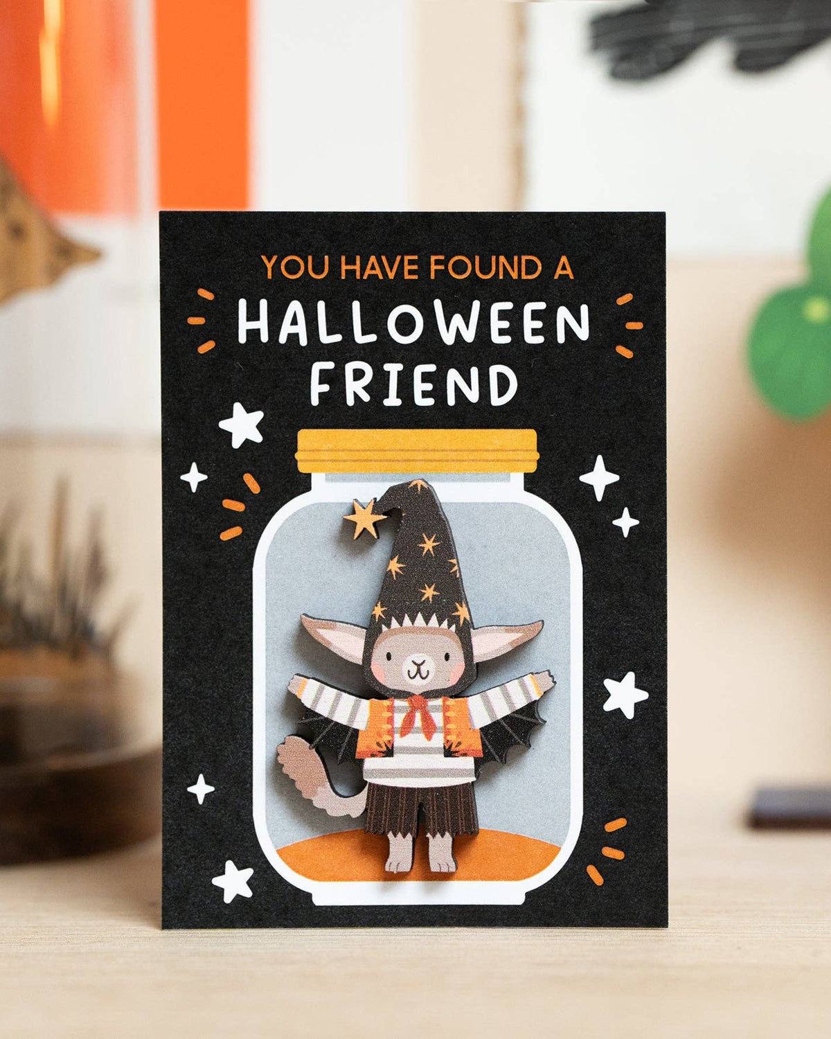 A Halloween pin on backing card standing upright on a tabletop, featuring a black background with the text 'YOU HAVE FOUND A HALLOWEEN FRIEND' and a cute cat dressed as a witch pin, inside a glass jar illustration.