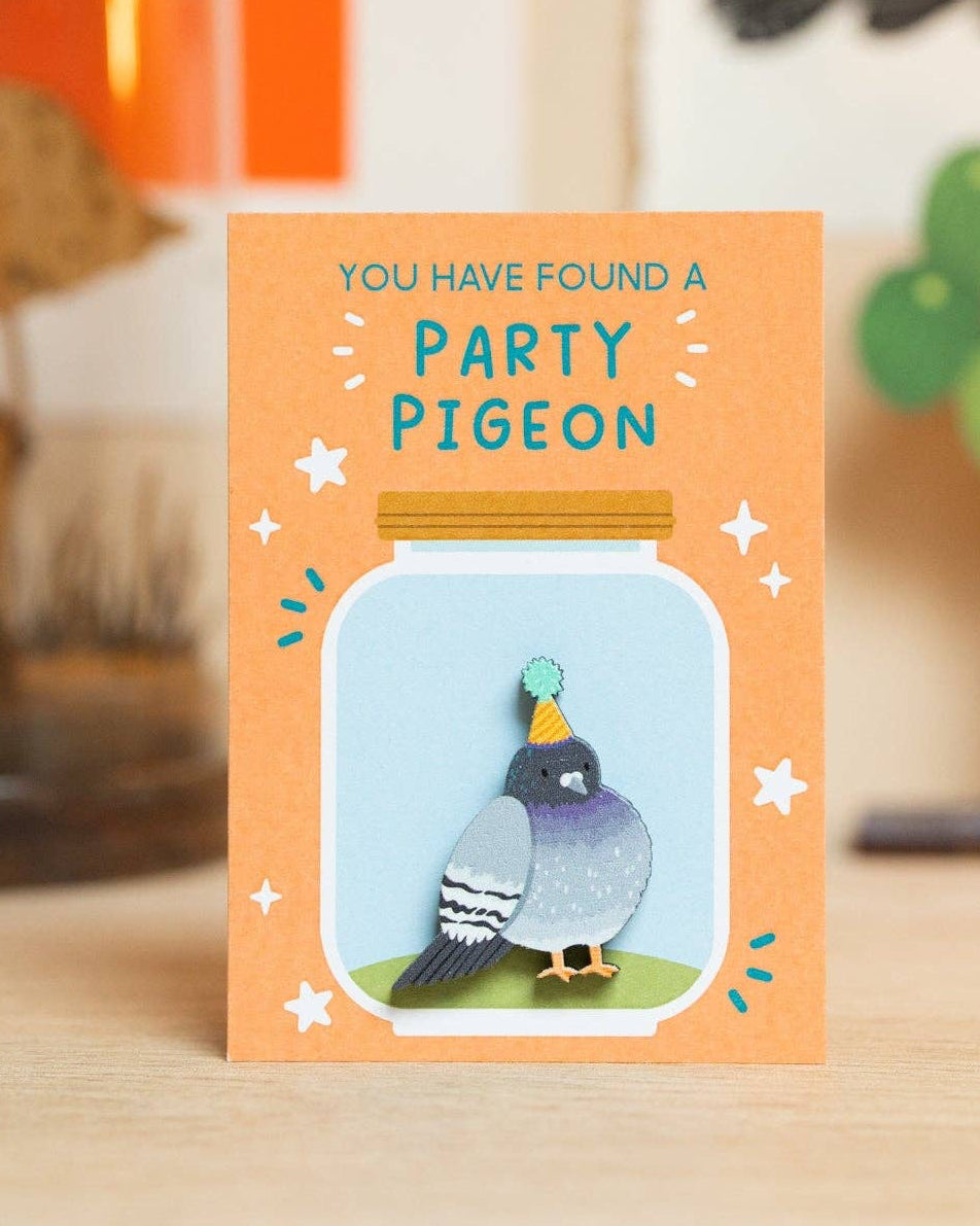 Wooden pigeon pin on a backing card with a peach-orange border, on a light wood surface. The card features the text 'YOU HAVE FOUND A PARTY PIGEON' above a pigeon pin, made to look like it’s inside a glass jar.