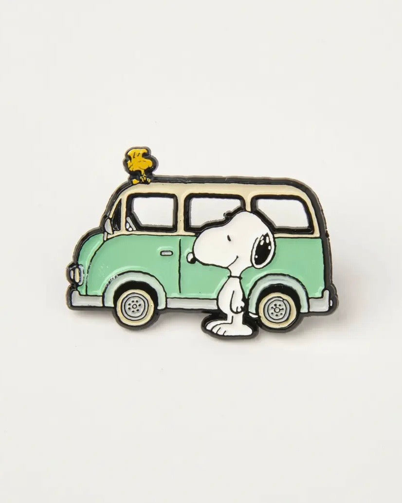 Enamel pin of Snoopy standing next to a vintage campervan on a light grey background