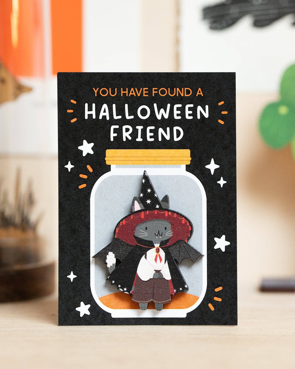 A Halloween pin on a backing card with the text 'YOU HAVE FOUND A HALLOWEEN FRIEND' and an illustration of a grey cat dressed as a witch inside a glass jar, displayed on a tabletop with a softly blurred home background.