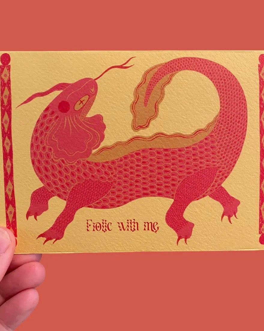 Card with red dragon illustration and 'Frolic with me' text on a gold background, held against a red background.